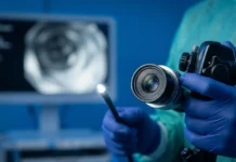 Cambios en las heces por cáncer de colon: 4 Alertas Vitales Fotografía médica macro en primer plano fotorrealista de un endoscopio moderno, con el lente de fibra óptica brillando intensamente, sostenido con firmeza por manos enguantadas quirúrgicas de nitrilo azul oscuro, en un entorno clínico esterilizado de fondo azul quirúrgico desenfocado.