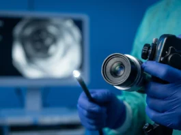 Fotografía médica macro en primer plano fotorrealista de un endoscopio moderno, con el lente de fibra óptica brillando intensamente, sostenido con firmeza por manos enguantadas quirúrgicas de nitrilo azul oscuro, en un entorno clínico esterilizado de fondo azul quirúrgico desenfocado.