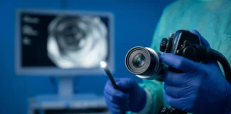 Cambios en las heces por cáncer de colon: 4 Alertas Vitales Fotografía médica macro en primer plano fotorrealista de un endoscopio moderno, con el lente de fibra óptica brillando intensamente, sostenido con firmeza por manos enguantadas quirúrgicas de nitrilo azul oscuro, en un entorno clínico esterilizado de fondo azul quirúrgico desenfocado.