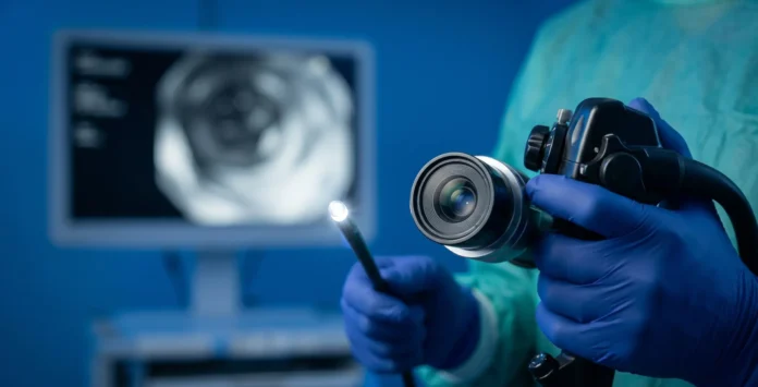 Fotografía médica macro en primer plano fotorrealista de un endoscopio moderno, con el lente de fibra óptica brillando intensamente, sostenido con firmeza por manos enguantadas quirúrgicas de nitrilo azul oscuro, en un entorno clínico esterilizado de fondo azul quirúrgico desenfocado.